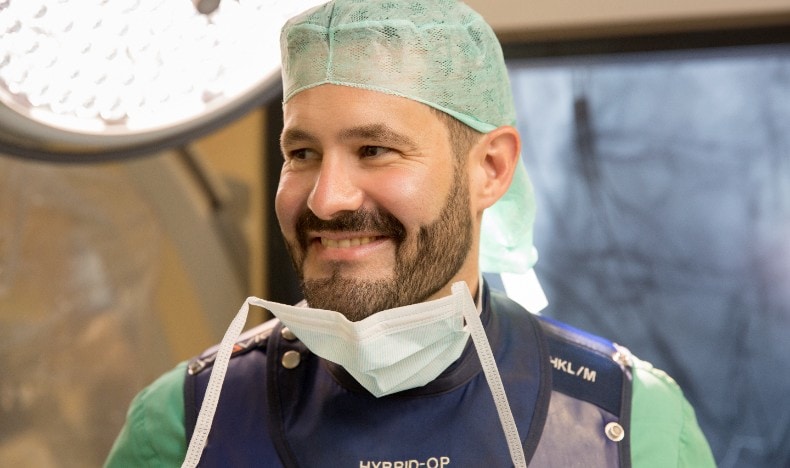 A smiling healthcare professional wearing surgical attire, including a green cap, protective vest labeled &ldquo;HYBRID-OP,&rdquo; and a surgical mask lowered under the chin. Medical equipment and a softly lit operating room environment are visible in the background, conveying a relaxed moment during a clinical procedure.
