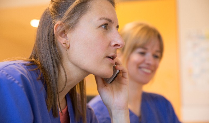 A close-up side profile of a healthcare professional wearing blue medical scrubs, holding a phone to her ear and listening attentively. Her hair is pulled back, and she appears focused. In the softly blurred background, another person in blue scrubs smiles, suggesting a calm, professional healthcare environment with warm indoor lighting.