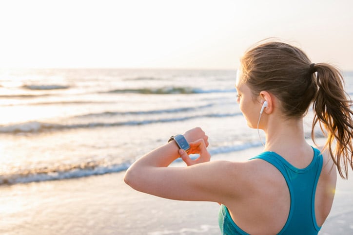 Over the shoulder view of mid adult woman by ocean wearing earbuds looking at wrist watch